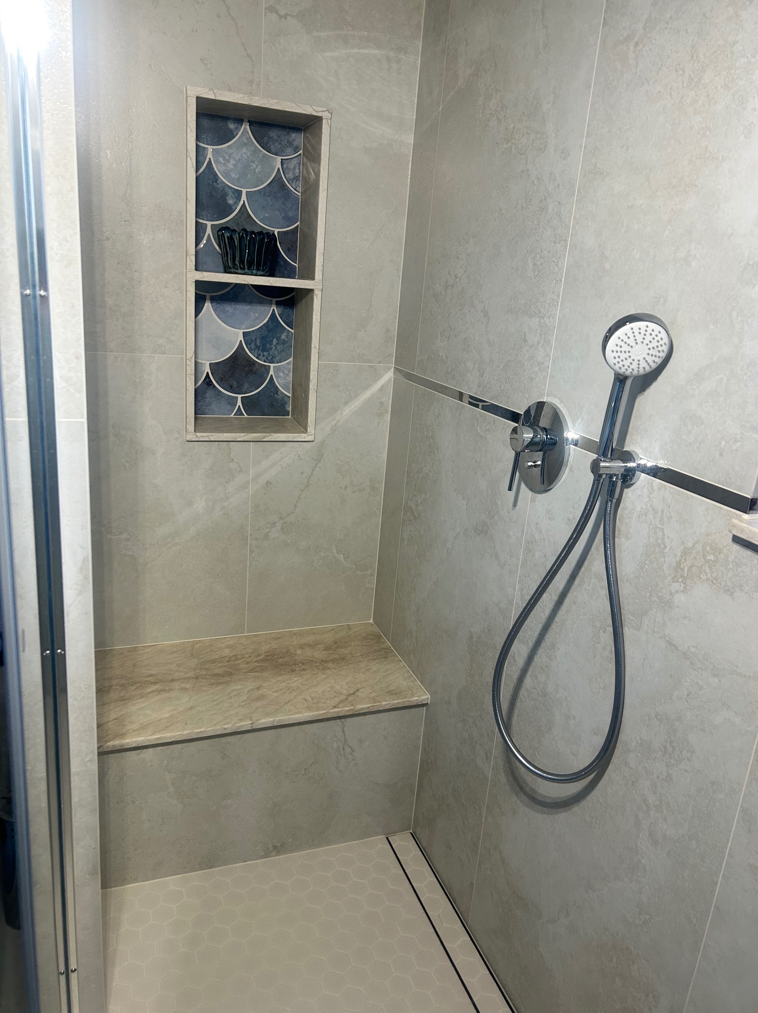 Stylowa kabina prysznicowa z półką wnękową wykończoną mozaiką w odcieniach niebieskiego. Jasne płytki imitujące kamień, siedzisko i heksagonalna podłoga.