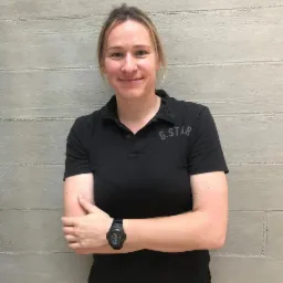 Portret uśmiechniętej kobiety w czarnej koszulce polo G-Star i czarnym zegarku na tle szarej ściany z poziomymi liniami.