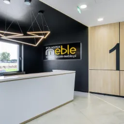 Nowoczesne biuro: biała recepcja z podświetleniem LED, czarne ściany z logo firmy meblarskiej, drewniane panele ścienne z numerem '1', geometryczna lampa wisząca.
