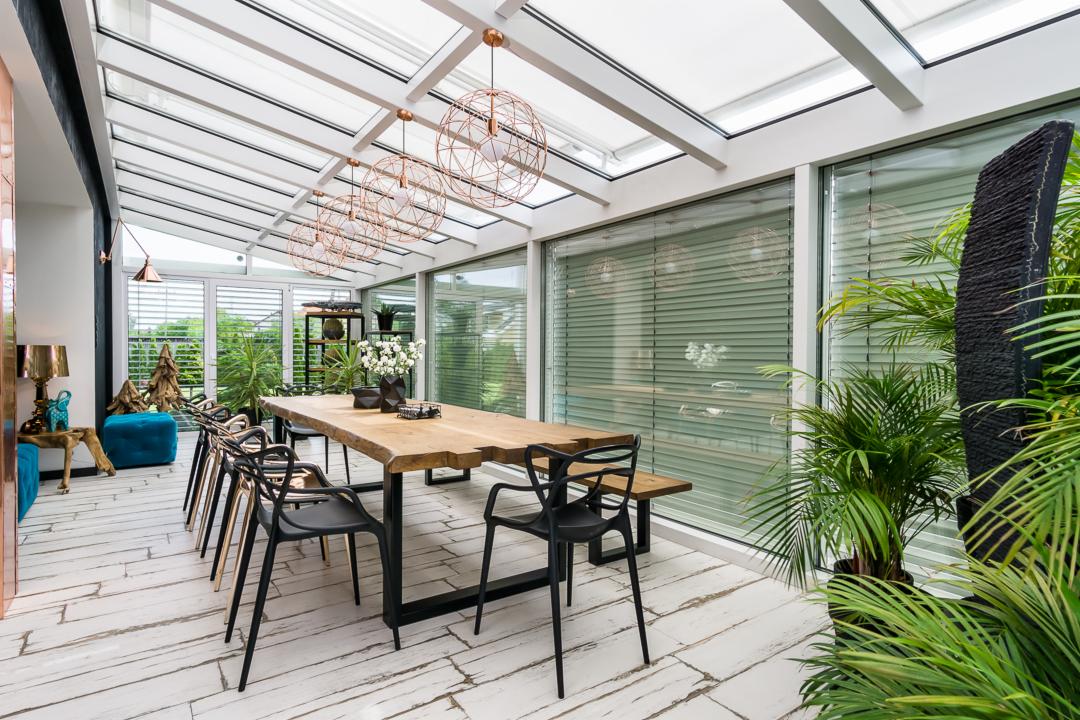 Jasna jadalnia w stylu nowoczesnym z przeszklonym dachem, drewnianym stołem na metalowych nogach, czarnymi krzesłami i dekoracyjnymi lampami w kształcie kuli