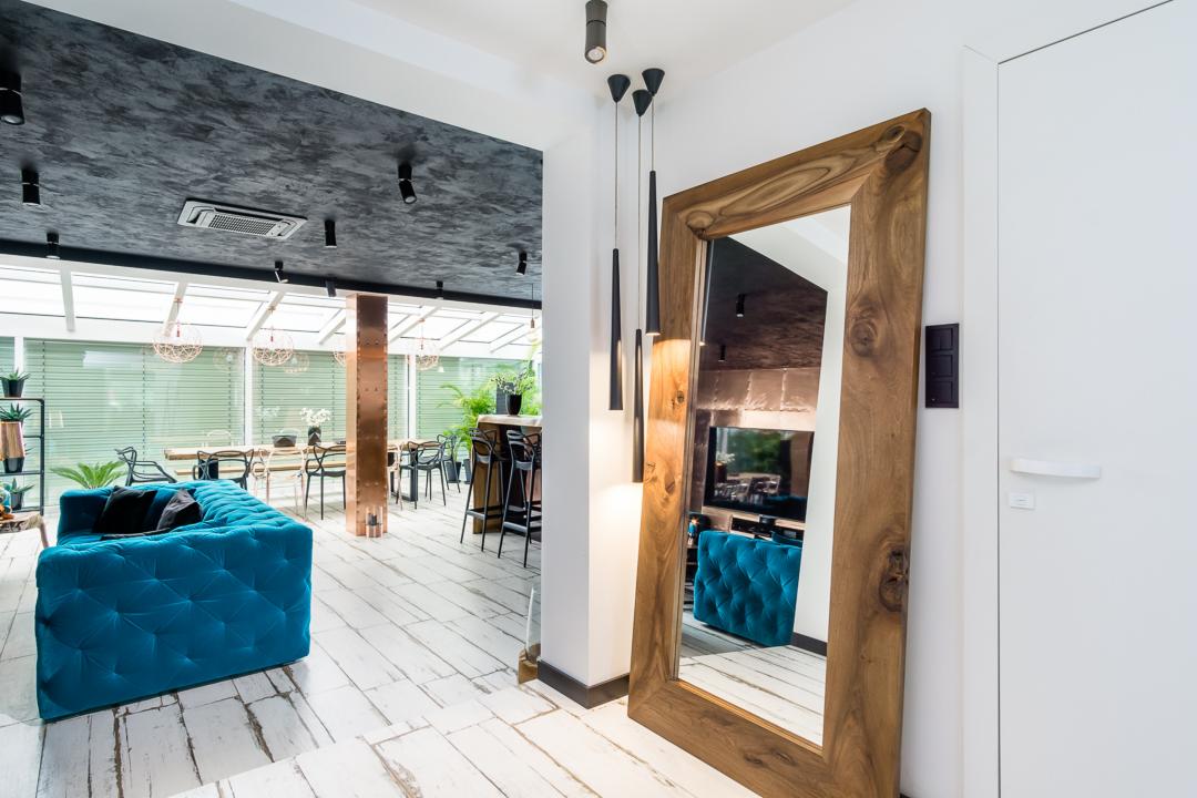 Nowoczesne wnętrze z turkusową sofą, dużym drewnianym lustrem i białymi deskami podłogowymi, widok na przeszklony salon z jadalnią w tle.