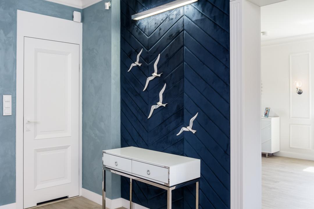 Nowoczesny przedpokój z granatową ścianą z paneli w jodełkę, dekoracją w kształcie mew, białą konsolą na chromowanych nogach oraz błękitną ścianą z efektem dekoracyjnym.
