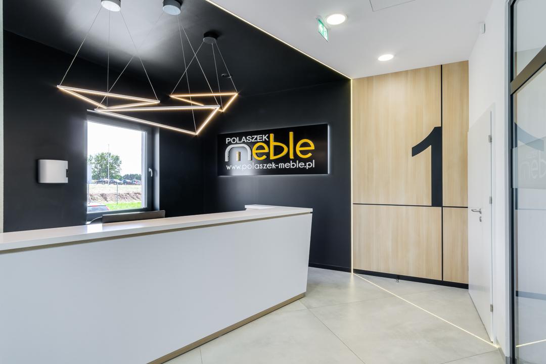 Nowoczesne biuro: biała recepcja z podświetleniem LED, czarne ściany z logo firmy meblarskiej, drewniane panele ścienne z numerem '1', geometryczna lampa wisząca.