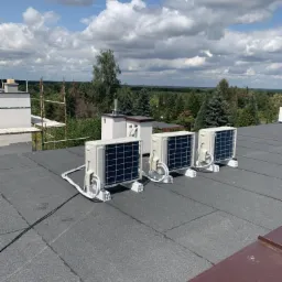 Cztery jednostki zewnętrzne klimatyzacji zamontowane na płaskim, pokrytym papą dachu budynku z widokiem na las i błękitne niebo z chmurami.