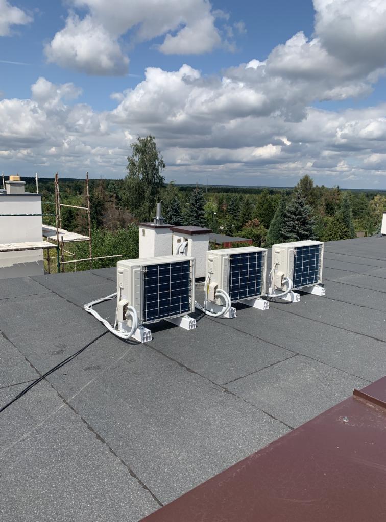 Cztery jednostki zewnętrzne klimatyzacji zamontowane na płaskim, pokrytym papą dachu budynku z widokiem na las i błękitne niebo z chmurami.