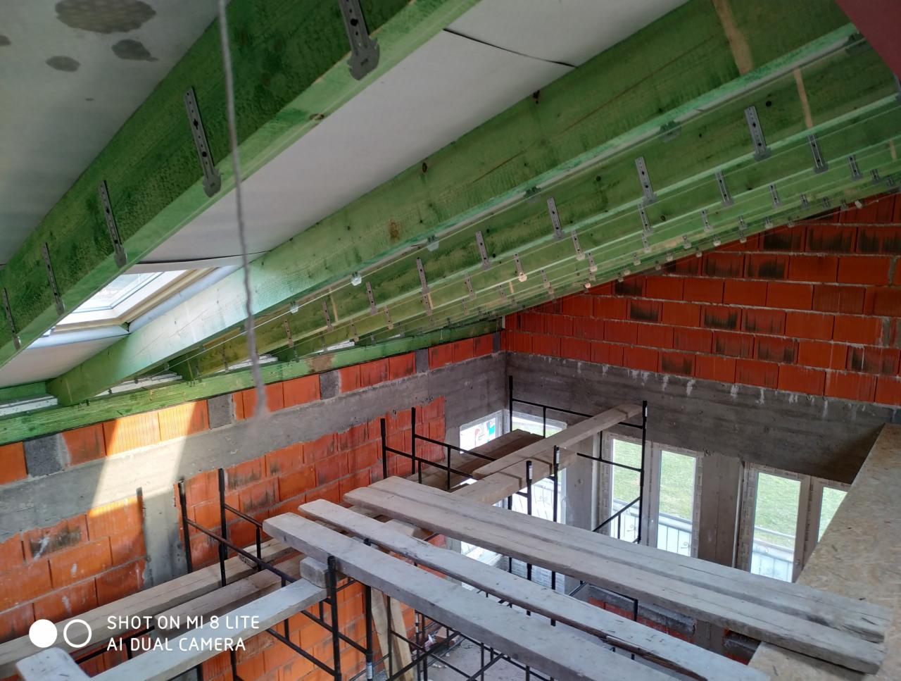 Poddasze w trakcie prac wykończeniowych: widoczne zielone drewniane belki stropowe z metalowymi mocowaniami, ceglana ściana i fragment rusztowania z drewnianymi deskami, okno dachowe.