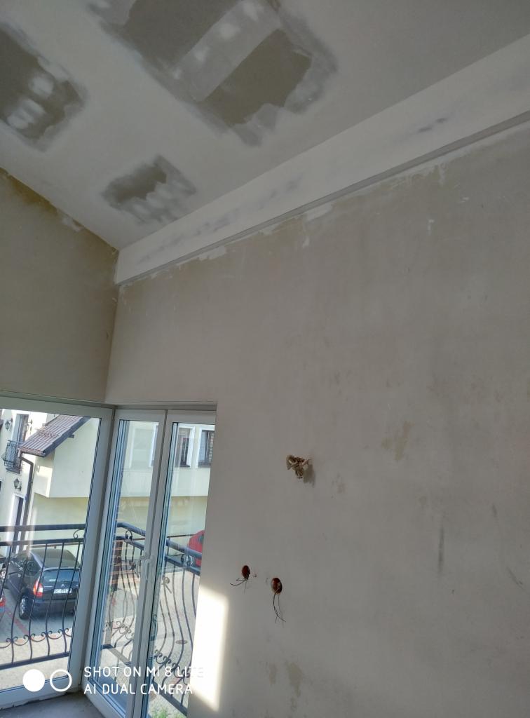 Pokój w trakcie remontu: widoczne niedoskonałości na suficie, częściowo wykończona ściana z wystającymi przewodami elektrycznymi i zamontowaną listwą przysufitową. Przez otwarte drzwi balkonowe...