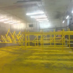 Żółte, metalowe konstrukcje zawieszone w komorze lakierniczej po malowaniu proszkowym, z widocznym pyłem farby na podłodze i oświetleniem przemysłowym.