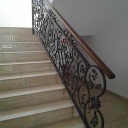 Nie Podano - Kuta balustrada schodowa z dekoracyjnymi elementami roślinnymi, drewniana poręcz, beżowe płytki na schodach i podłodze.