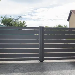 Nowoczesna, pozioma brama wjazdowa w kolorze antracytowym, zamontowana na betonowych słupkach z domofonem i lampą ostrzegawczą, na tle domu z brązowym dachem.