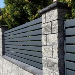Nowoczesne ogrodzenie z poziomych, szarych paneli metalowych osadzonych między kamiennymi słupkami i podmurówką, widok z chodnika.