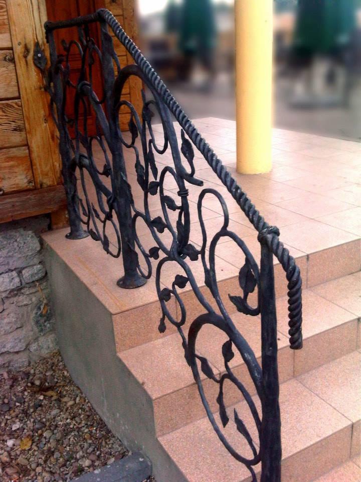 Kuta balustrada schodowa z motywem roślinnym, czarna, z grubą poręczą przypominającą linę, zamontowana przy wejściu do domu wyłożonym płytkami.