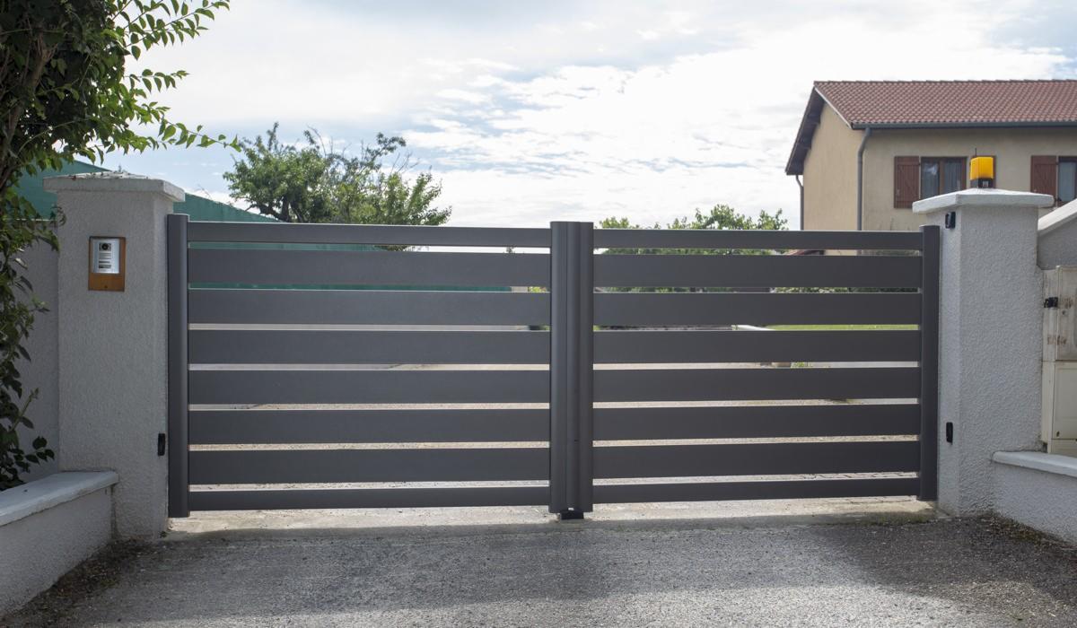 Nowoczesna, pozioma brama wjazdowa w kolorze antracytowym, zamontowana na betonowych słupkach z domofonem i lampą ostrzegawczą, na tle domu z brązowym dachem.