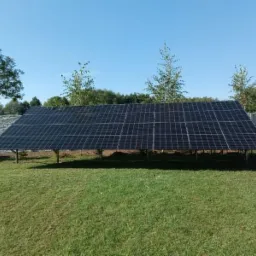 Instalacja paneli słonecznych na zielonym trawniku, widok z niskiej perspektywy. Szary, betonowy płot w tle. Błękitne niebo.