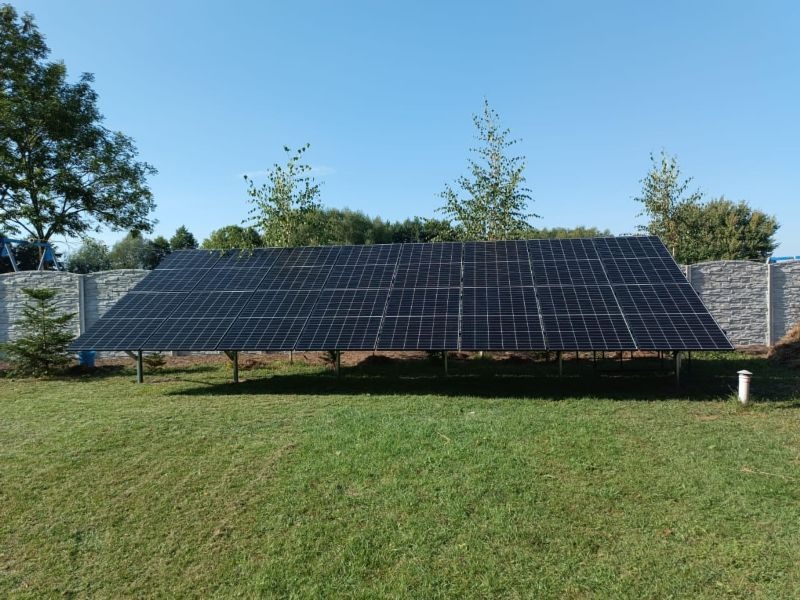 Instalacja paneli słonecznych na zielonym trawniku, widok z niskiej perspektywy. Szary, betonowy płot w tle. Błękitne niebo.