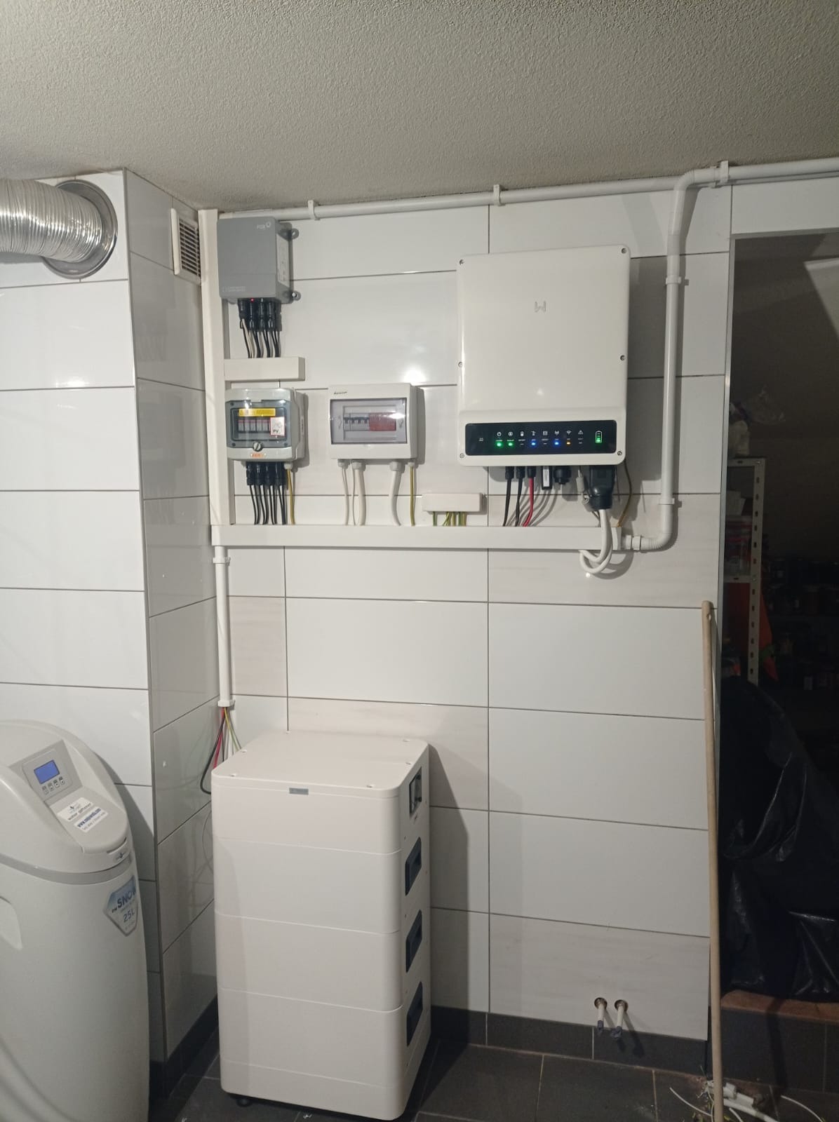 Biały magazyn energii z podłączonym inwerterem na ścianie z kafelkami. Widoczne zabezpieczenia, przewody i rury instalacyjne. Domowa instalacja w pomieszczeniu.