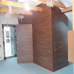 Wnętrze w trakcie wykończenia z widoczną drewnianą obudową kominka z otworami na gniazdka elektryczne, dopasowaną kolorystycznie drewnianą ścianą oraz otwartymi drzwiami wewnętrznymi z drewnianym...