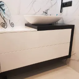 Nowoczesna szafka łazienkowa z białymi frontami szuflad i czarnym blatem pod umywalkę nablatową, uzupełniona o dekoracyjne lusterko kosmetyczne na metalowej podstawie, na tle marmurowych płytek...