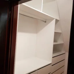 Wnętrze białej garderoby wnękowej z szufladami i półkami, widoczne z perspektywy wejścia, oświetlone punktowo.