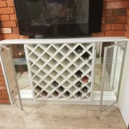 Biała szafka pod telewizor z ażurowym regałem na wino i przeszklonymi szafkami bocznymi, na tle ściany z czerwonej cegły.