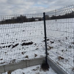 Kamil Woźniak - Fragment nowo zamontowanego, panelowego ogrodzenia z podmurówką betonową na zasypanym śniegiem terenie; widoczne łączenia paneli i słupków, w tle pole z resztkami śniegu i drzewa.