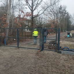 Kamil Woźniak - Montaż nowoczesnej, metalowej bramy ogrodzeniowej z siatki na posesji; widoczni dwaj pracownicy w trakcie instalacji, jeden w kamizelce odblaskowej, w tle drzewa i nowy dom.