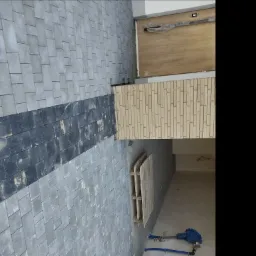 Szara elewacja budynku z elementami dekoracyjnymi z cegły i ciemnym pasem izolacji termicznej, widoczne niebieskie rury i narzędzia na podłodze wewnątrz budynku.