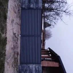 Nowoczesna, grafitowa brama wjazdowa z poziomymi panelami, osadzona na betonowych słupkach, na tle surowego stanu budynku z cegły i drzewa bez liści.