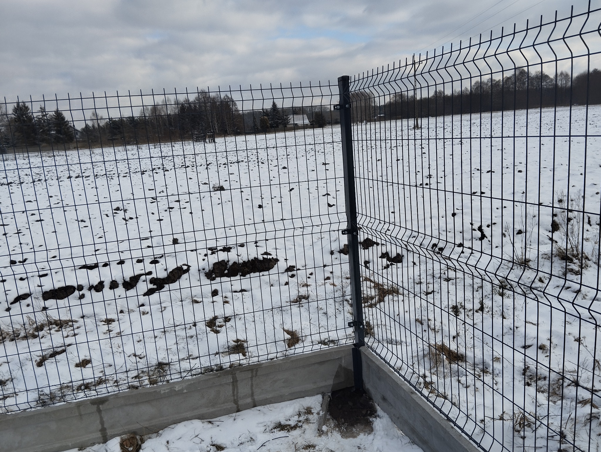Fragment nowo zamontowanego, panelowego ogrodzenia z podmurówką betonową na zasypanym śniegiem terenie; widoczne łączenia paneli i słupków, w tle pole z resztkami śniegu i drzewa.