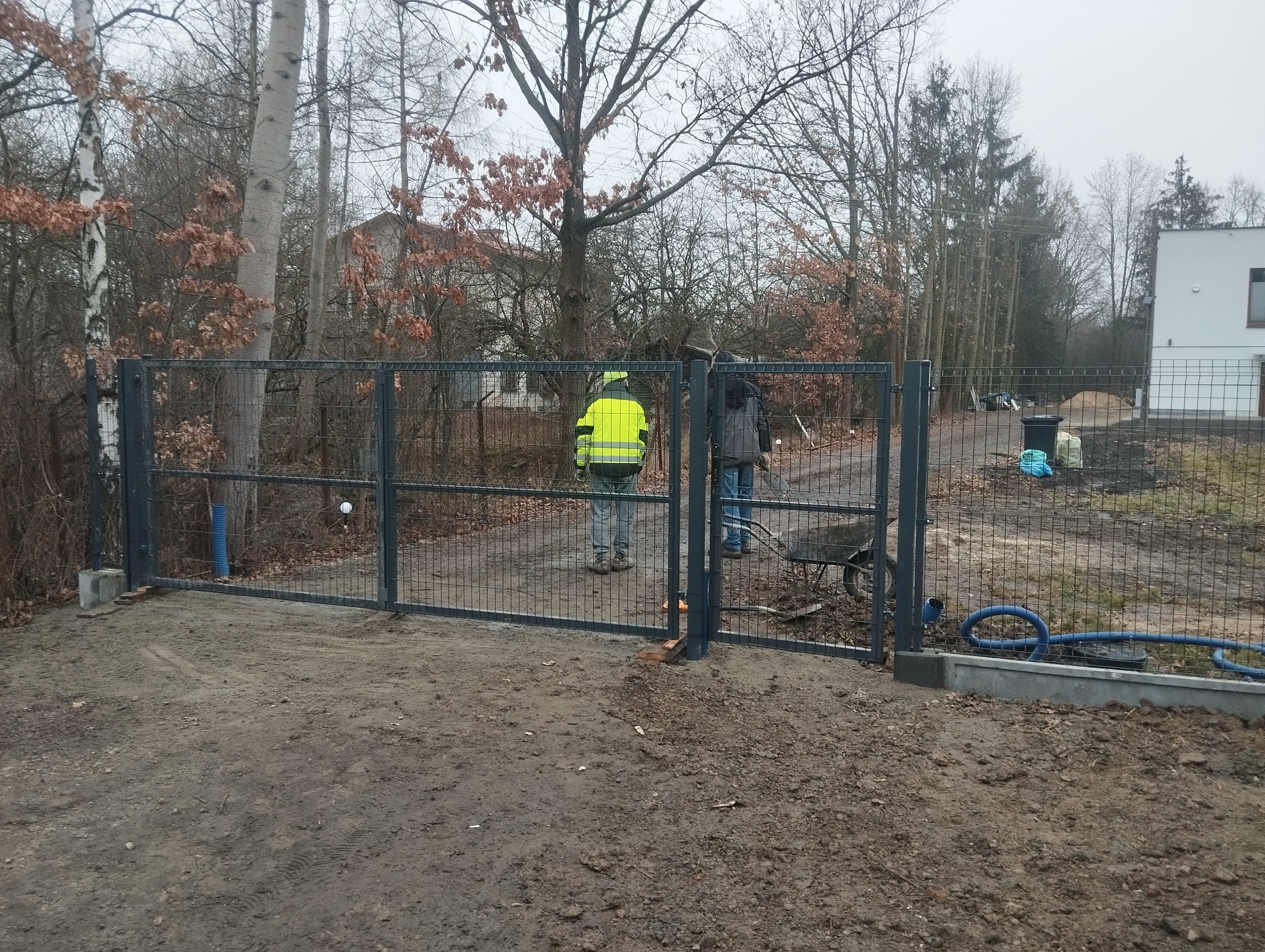 Montaż nowoczesnej, metalowej bramy ogrodzeniowej z siatki na posesji; widoczni dwaj pracownicy w trakcie instalacji, jeden w kamizelce odblaskowej, w tle drzewa i nowy dom.