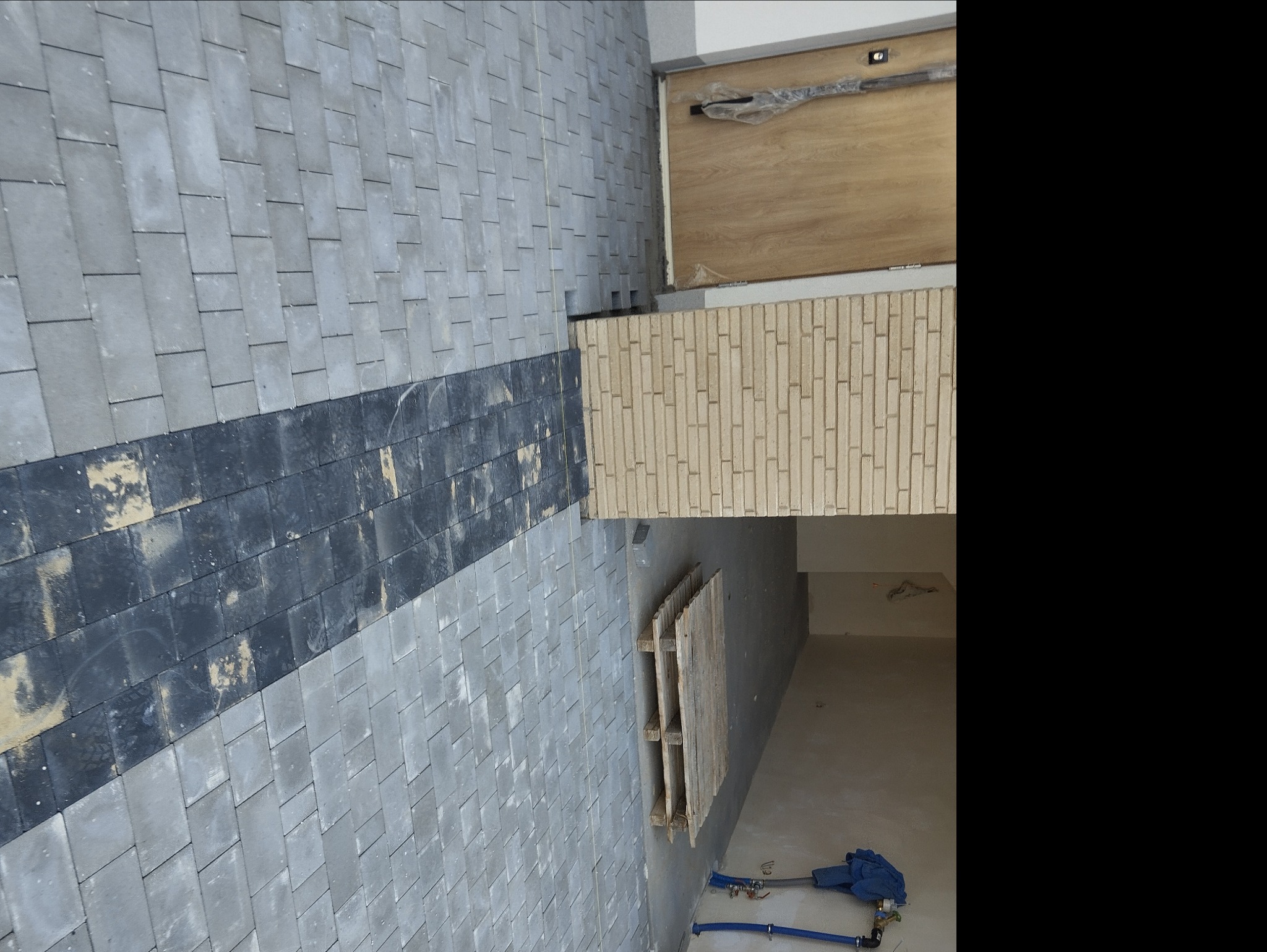 Szara elewacja budynku z elementami dekoracyjnymi z cegły i ciemnym pasem izolacji termicznej, widoczne niebieskie rury i narzędzia na podłodze wewnątrz budynku.