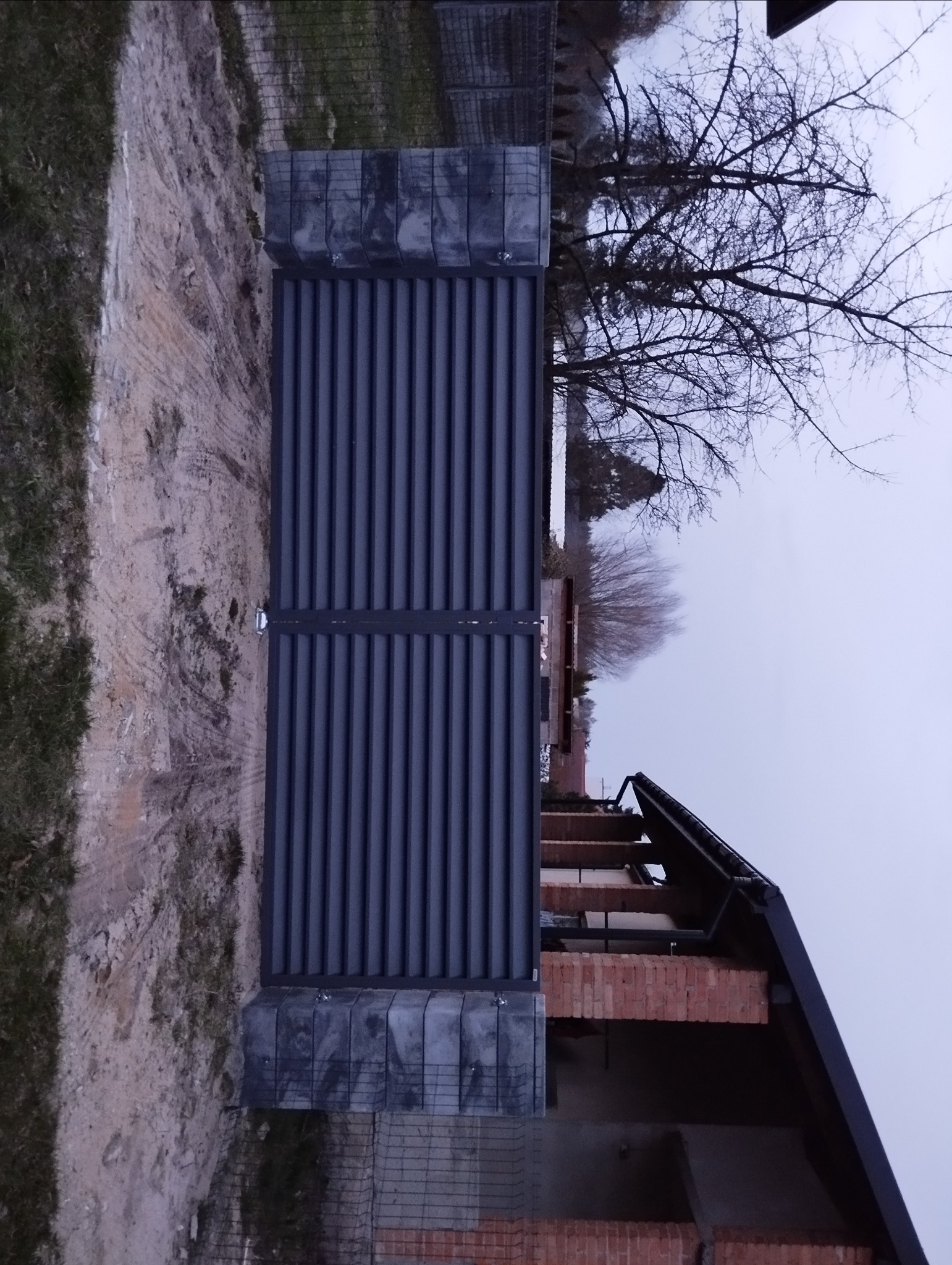 Nowoczesna, grafitowa brama wjazdowa z poziomymi panelami, osadzona na betonowych słupkach, na tle surowego stanu budynku z cegły i drzewa bez liści.