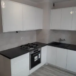 Nowoczesna, biała zabudowa kuchenna z ciemnym blatem, płyta gazowa i piekarnik w zabudowie, jasne płytki na ścianie, widok na umywalkę i baterię.