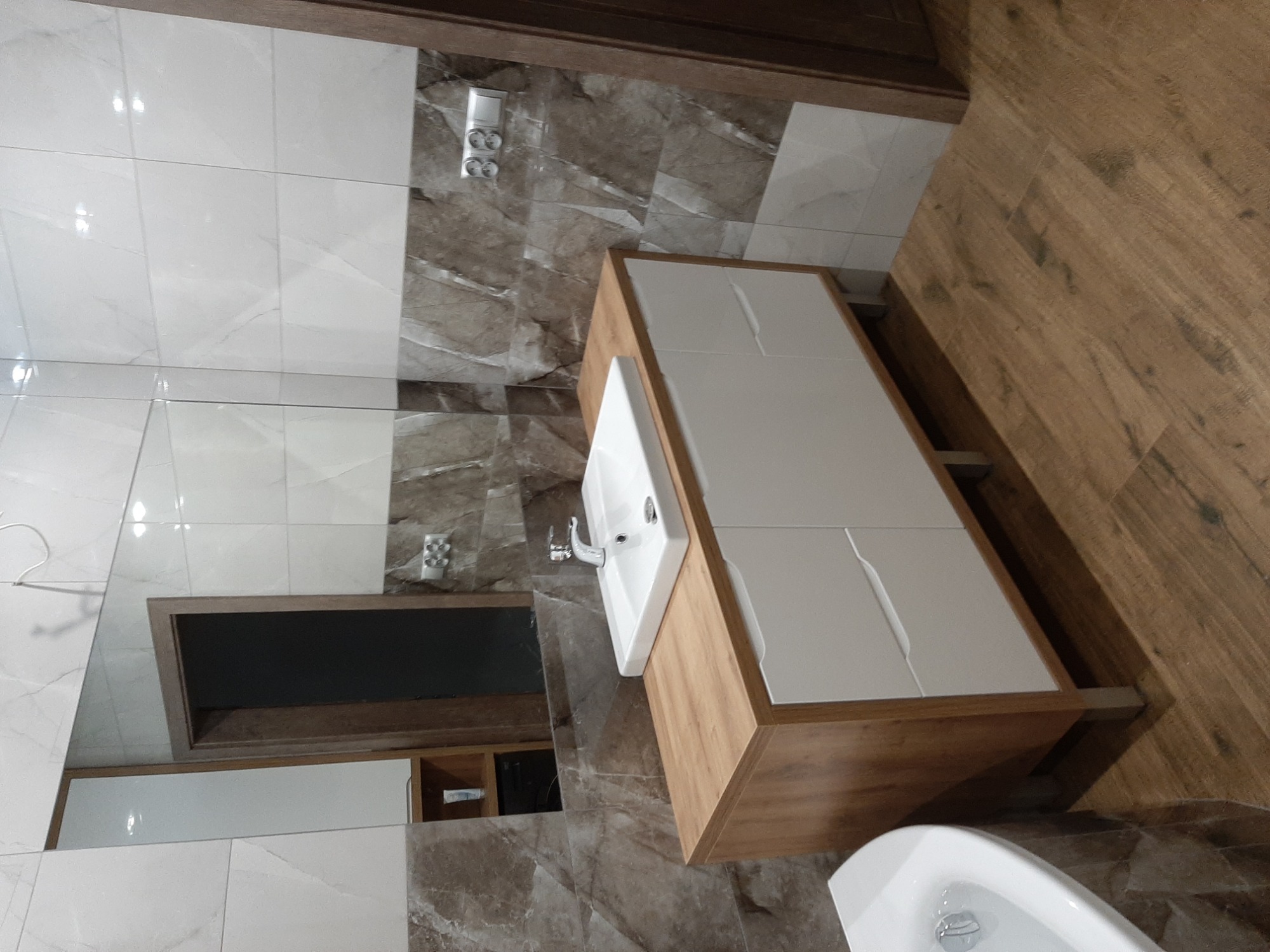 Nowoczesna łazienka z szafką pod umywalkę z drewnianym blatem i białymi frontami, marmurowe płytki na ścianach, drewnopodobna podłoga, widoczne odbicie lustra.