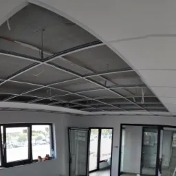 Montaż sufitu podwieszanego z płyt kartonowo-gipsowych w pomieszczeniu biurowym, widoczne metalowe profile konstrukcyjne i częściowo zamontowane płyty, instalacja wentylacyjna pod sufitem, okna...