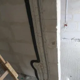 Wiązka przewodów elektrycznych, czarnych i szarych, przymocowana do surowej ściany z betonu komórkowego za pomocą zaprawy, z widocznym fragmentem drabiny malarskiej.