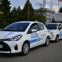 Flota trzech białych samochodów z niebieskimi akcentami i logo Primo Getin Auto Partner, zaparkowana na brukowanej ulicy przed budynkiem i billboardem reklamowym.