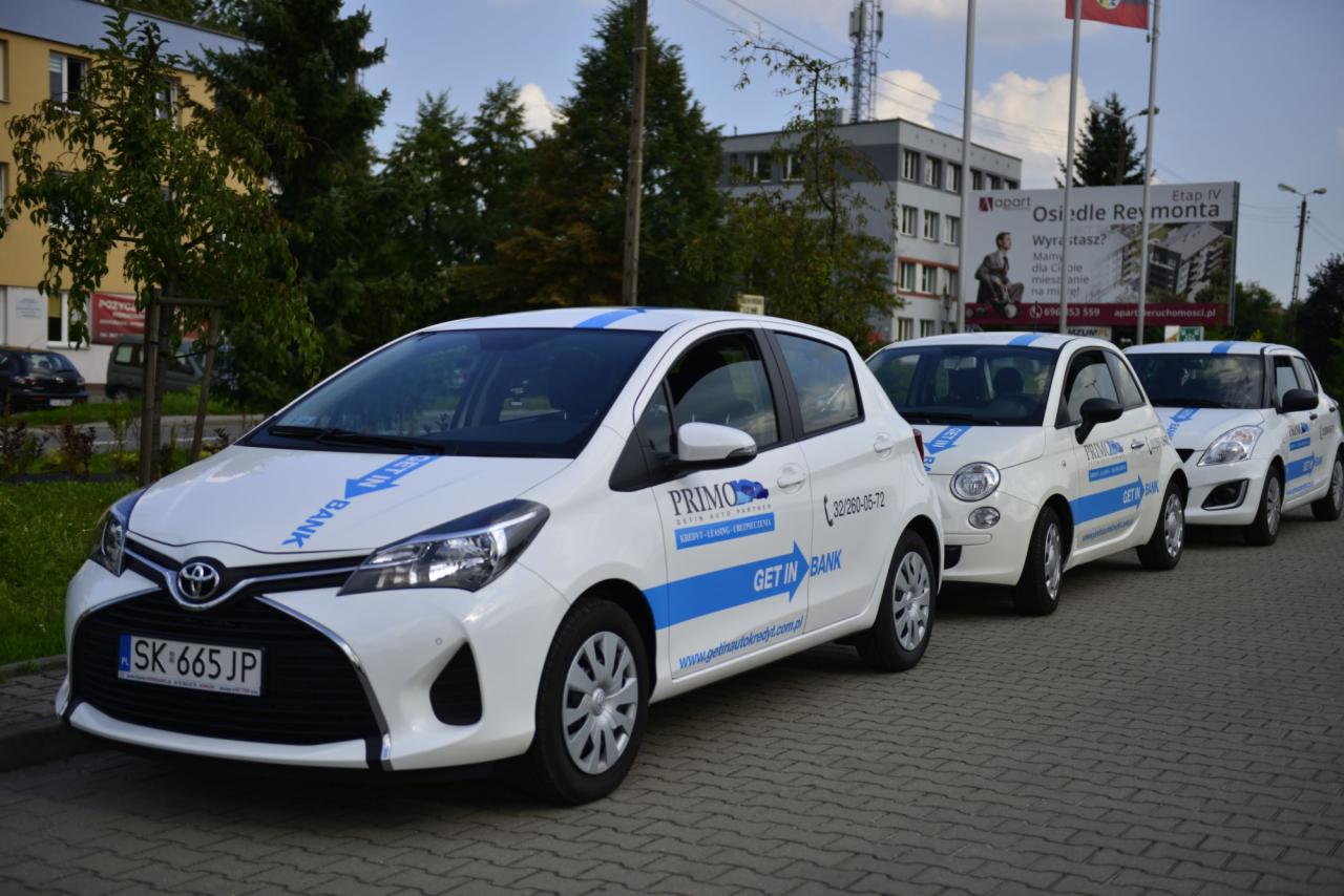 Flota trzech białych samochodów z niebieskimi akcentami i logo Primo Getin Auto Partner, zaparkowana na brukowanej ulicy przed budynkiem i billboardem reklamowym.