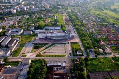 Panoramiczny widok z lotu ptaka na miasto z dużym budynkiem o zakrzywionej fasadzie, otoczonym zielenią i parkingami, w słoneczny dzień.