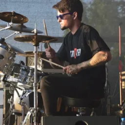 Perkusista z tatuażami gra na perkusji Pearl w okularach przeciwsłonecznych na scenie z nagłośnieniem TIBO w tle.