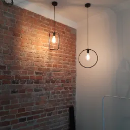 Dwie nowoczesne lampy wiszące z żarówkami Edisona, jedna w czarnej kwadratowej ramie, druga w okrągłej, na tle ceglanej ściany i białej ściany z listwą przysufitową. Widoczny zwój kabla...