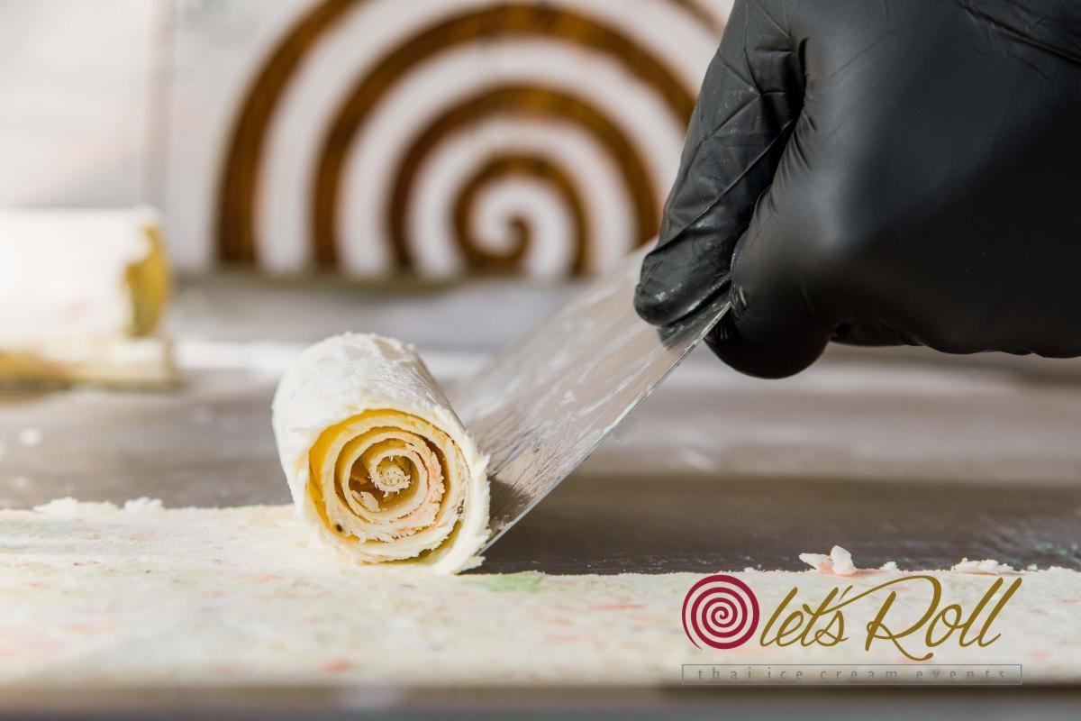 Przygotowywanie lodów tajskich: ręka w czarnej rękawiczce formuje rolkę lodową za pomocą metalowej szpatułki na chłodzącej płycie, w tle widoczny wzór spirali z polewy.