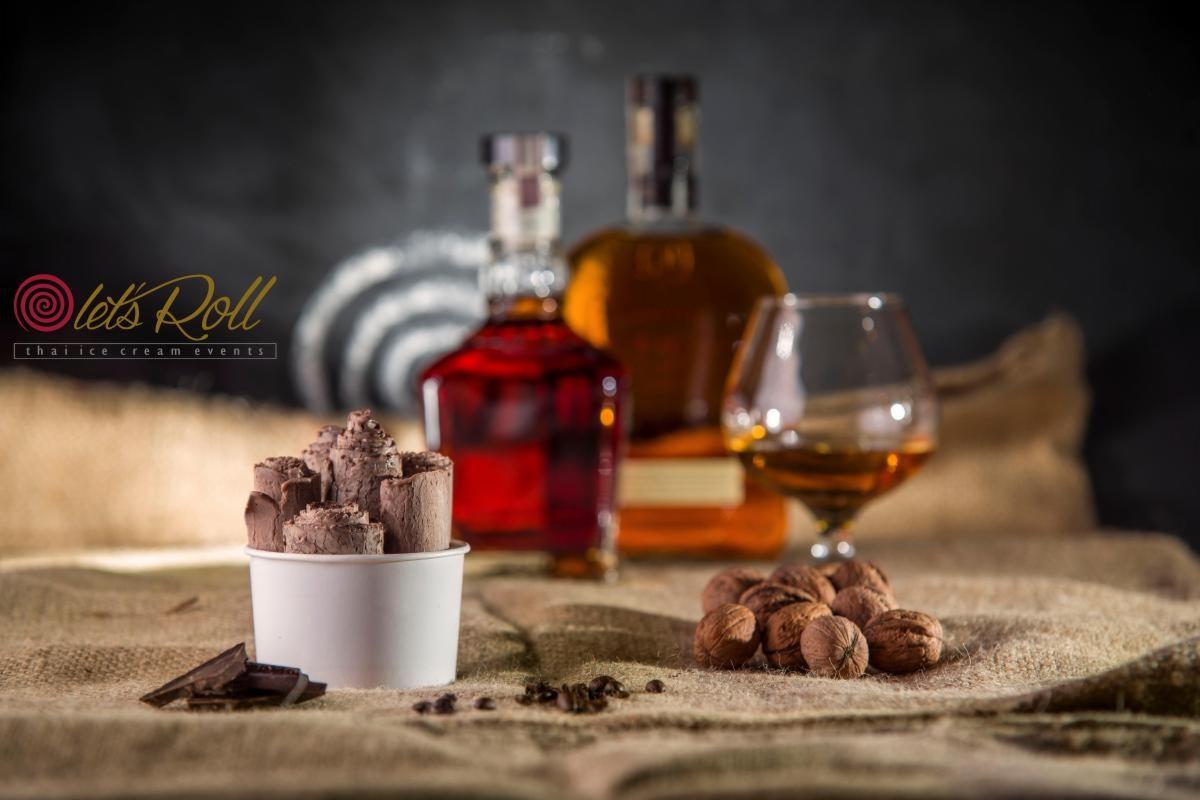 Tajskie lody czekoladowe w białym kubku, kawałki czekolady obok, w tle butelki alkoholu i orzechy włoskie na jutowym materiale.