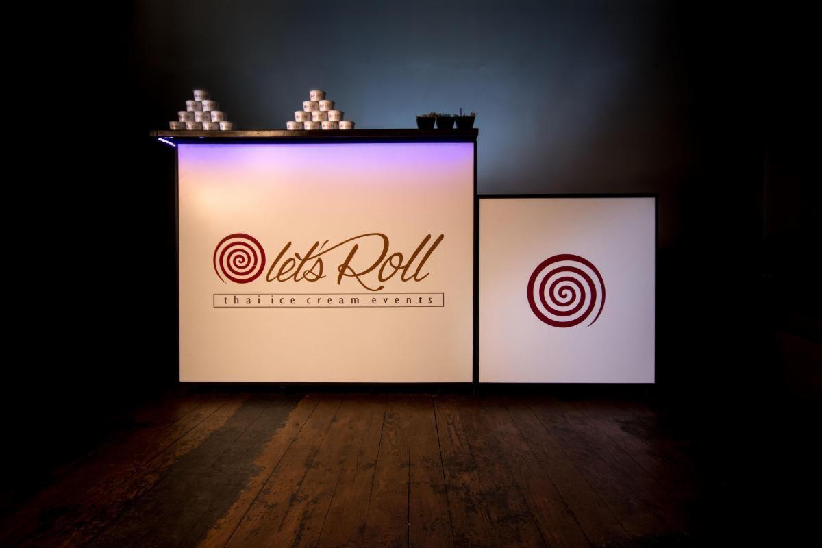 Mobilny bar z podświetleniem LED, logo 'Let's Roll Thai Ice Cream Events', piramidy z kubeczków na górnej półce, ciemne tło i drewniana podłoga.