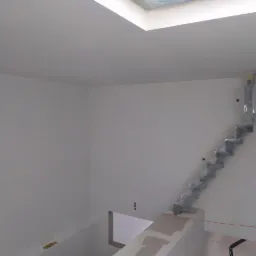 Wnętrze po malowaniu na biało, widoczne zabezpieczone folią schody z metalowymi stopniami, sufit z oknem dachowym, ściany przygotowane do dalszych prac wykończeniowych, widoczne niedoskonałości...