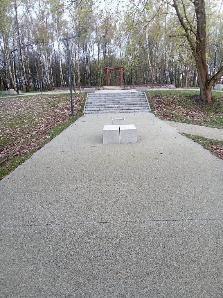 Parkowa ścieżka z dwoma betonowymi blokami prowadząca do schodów i czerwonej bramy torii na tle drzew.