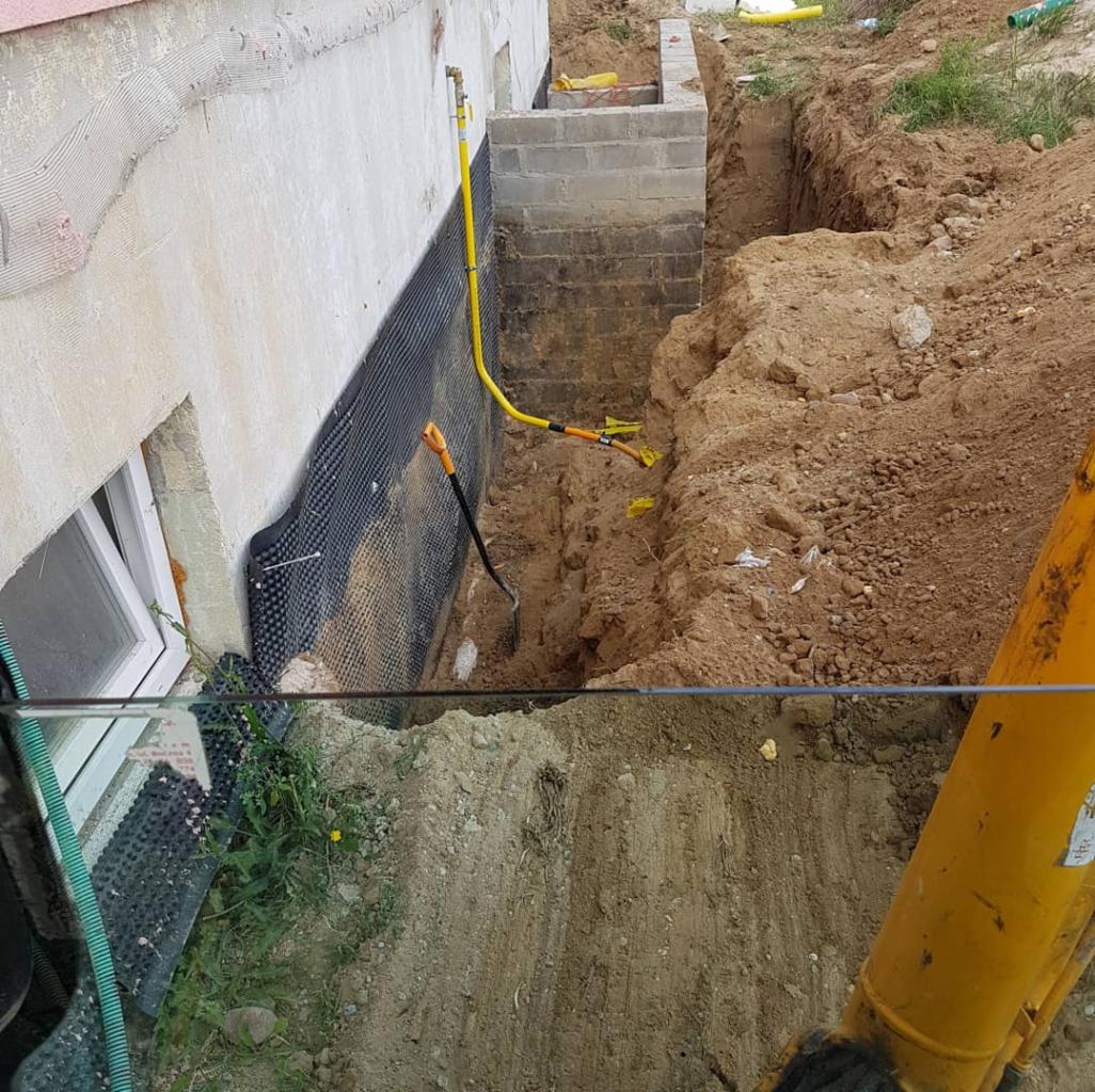 Wykop w ziemi wzdłuż fundamentów budynku, zabezpieczonych folią hydroizolacyjną i żółtą rurą gazową. W wykopie widoczna szpadel i fragment koparki.