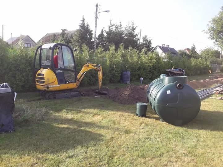 Żółta mini koparka New Holland wykonuje wykop obok zielonego, plastikowego szamba na trawiastym terenie działki, w tle żywopłot i zabudowa jednorodzinna.