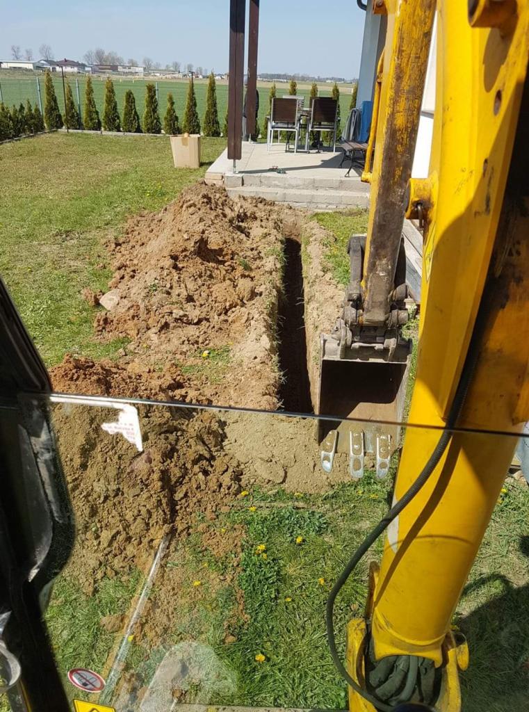 Wąski wykop w trawniku, widziany z kabiny żółtej koparki, z domem w tle i rzędem tuj.
