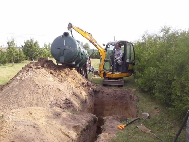 Żółta mini koparka umieszcza zielony, podłużny zbiornik w wykopie; widoczny operator, hałda ziemi i narzędzia.